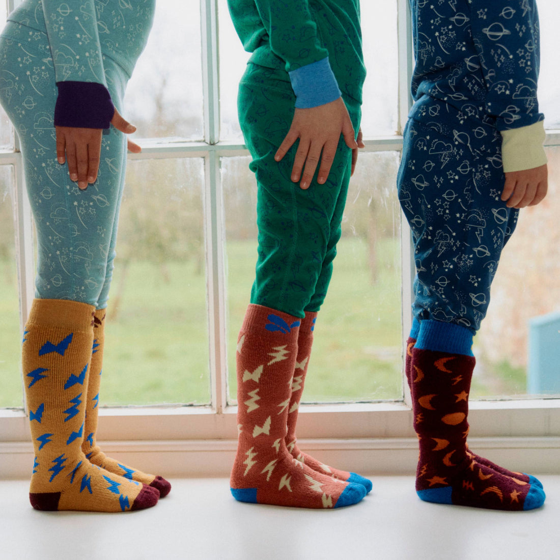 Three kids in space-themed Puddlestomper Twin Pack Socks, lined up by a window in their pyjamas. Each has unique sock and pyjama colours.