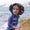 A child in Muddy Puddles waterproofs, wearing a hat and long-sleeved top, on a beach next to a rock, with the sea in the background.
