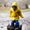 A child in Muddy Puddles waterproofs jumps in a muddy puddle, splashing mud everywhere. They are wearing a yellow coat and blue trousers.