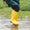 Muddy Puddles waterproofs: A child in a green coat and yellow wellies splashes in a puddle on a grassy field.