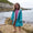 Muddy Puddles waterproofs: A happy girl with curly hair wears a blue raincoat patterned with clouds and raindrops on a rocky beach by the sea.
