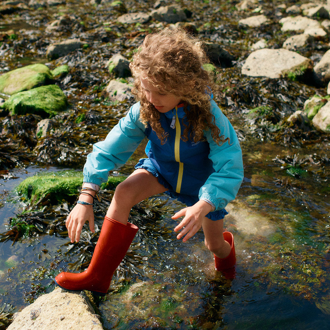 A girl with curly hair paddling in shallow water wearing 'Classic Wellies Red', jacket and shorts in a rocky, seaweed-filled area.