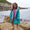 A young girl in a Muddy Puddlers! changing robe, smiling on a rocky beach, with the sea and coastline visible in the background.