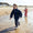 A young girl in a Muddy Puddles navy blue splash suit and wellies playing in the water on a beach, with another child behind.