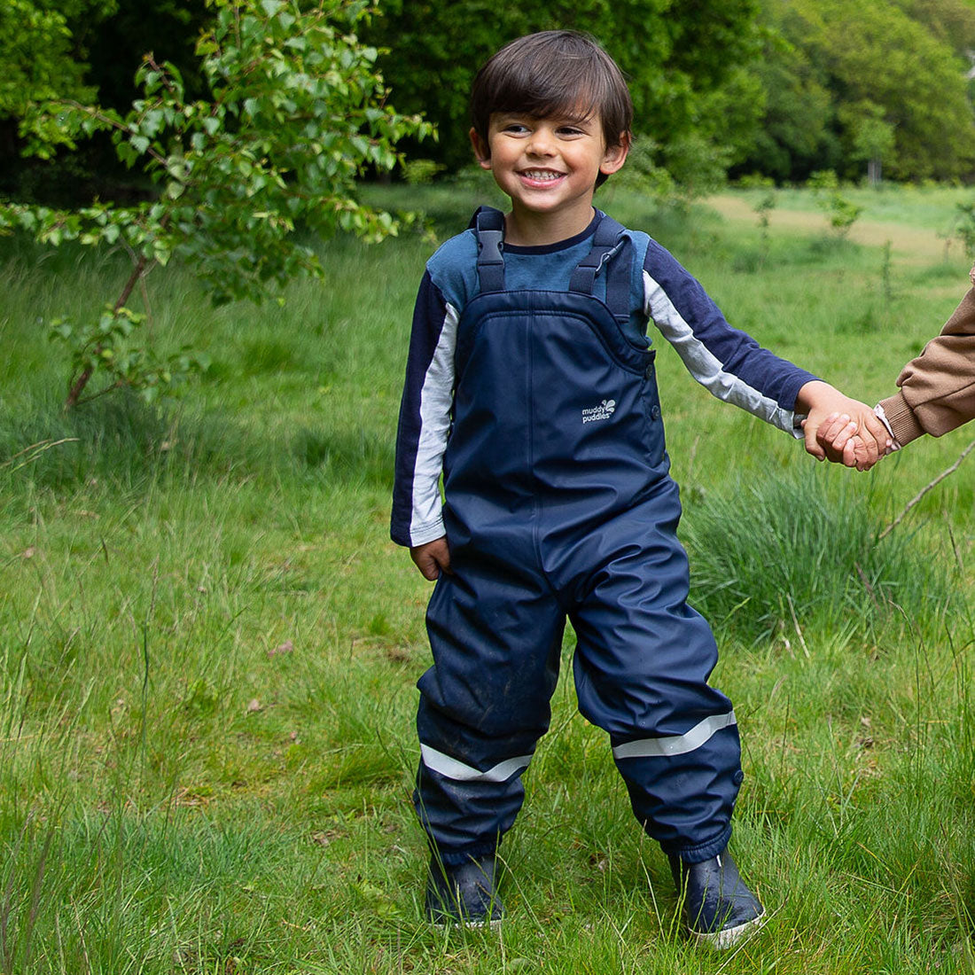 A young boy smiles in a field, wearing Puddleflex Waterproof Fleece Lined Dungarees in navy. He holds someone's hand, with trees behind.