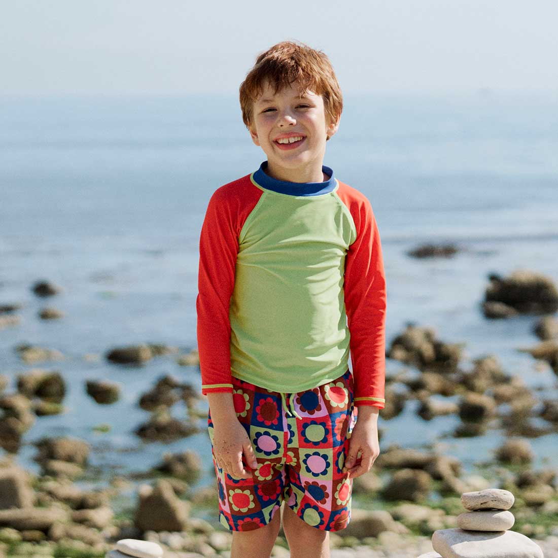 A boy wearing a UV Protective Rash Vest Multi Colourblock stands on a beach with rocks and the ocean in the background, smiling at the camera.