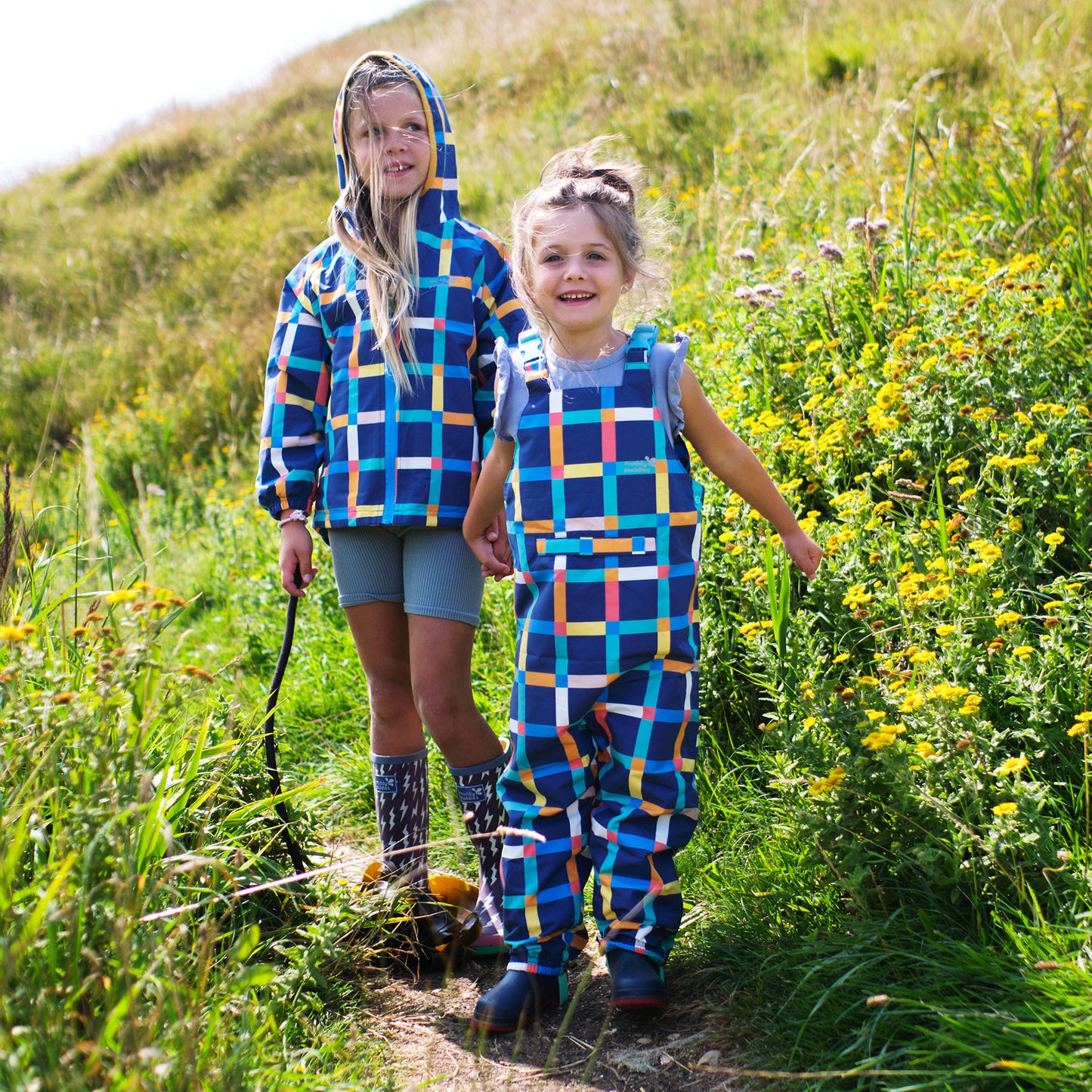 Two girls outdoors in a field. One wears an EcoLight Waterproof Dungarees Multi Check. Both are smiling and holding hands.