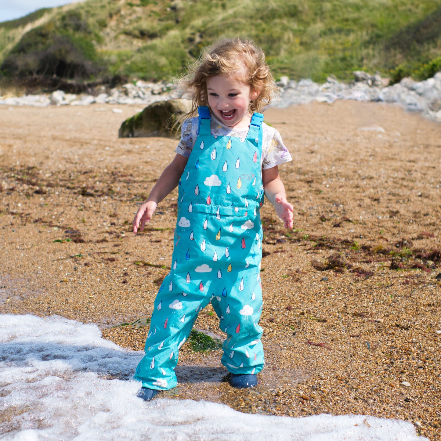 A young girl plays on a beach in EcoLight Waterproof Dungarees Green Raindrops. She is laughing as the water splashes around her feet.