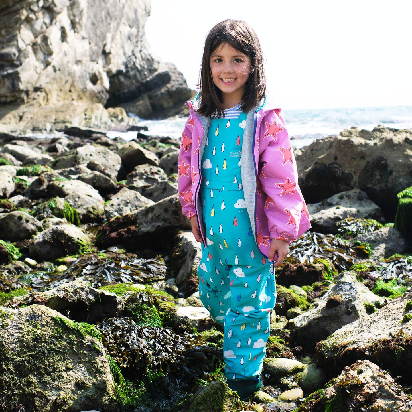 A girl stands on a rocky beach wearing EcoLight Waterproof Dungarees Green Raindrops with a pink starfish patterned jacket.