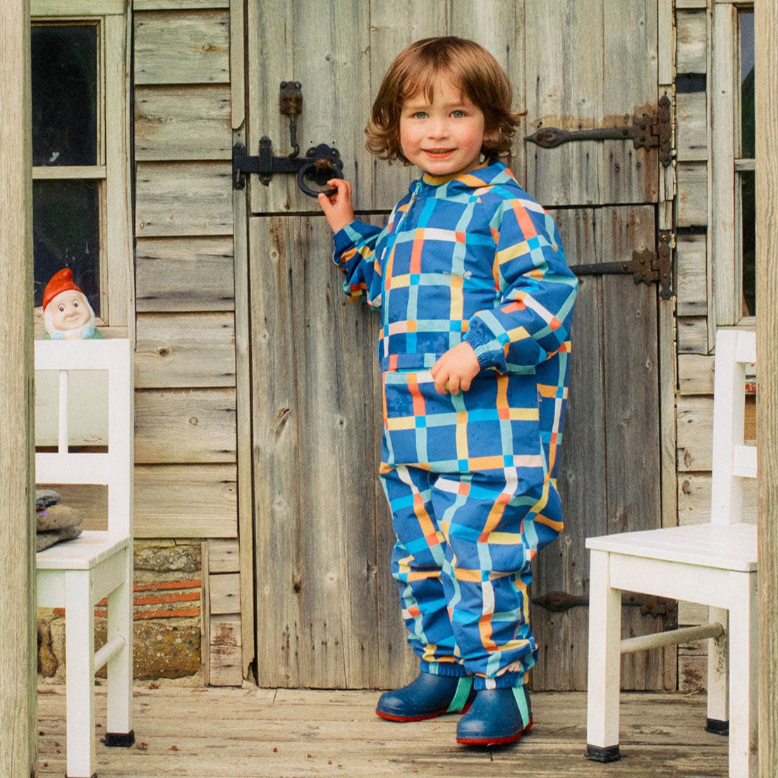 A young child smiles, wearing the EcoLight Lightweight Waterproof Puddle Suit Multi Check, standing on a wooden porch by a door.