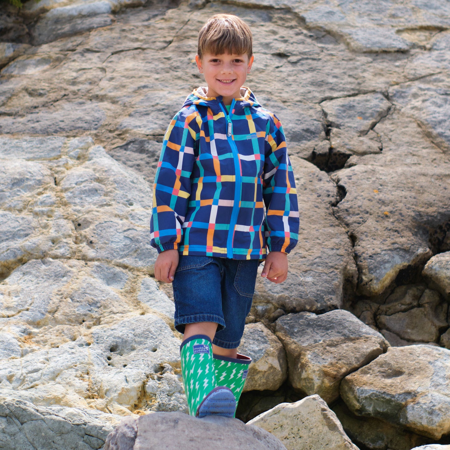 A boy stands on rocks wearing a Muddy Puddles EcoLight Lightweight Waterproof Jacket Multi Check, shorts and green wellies.