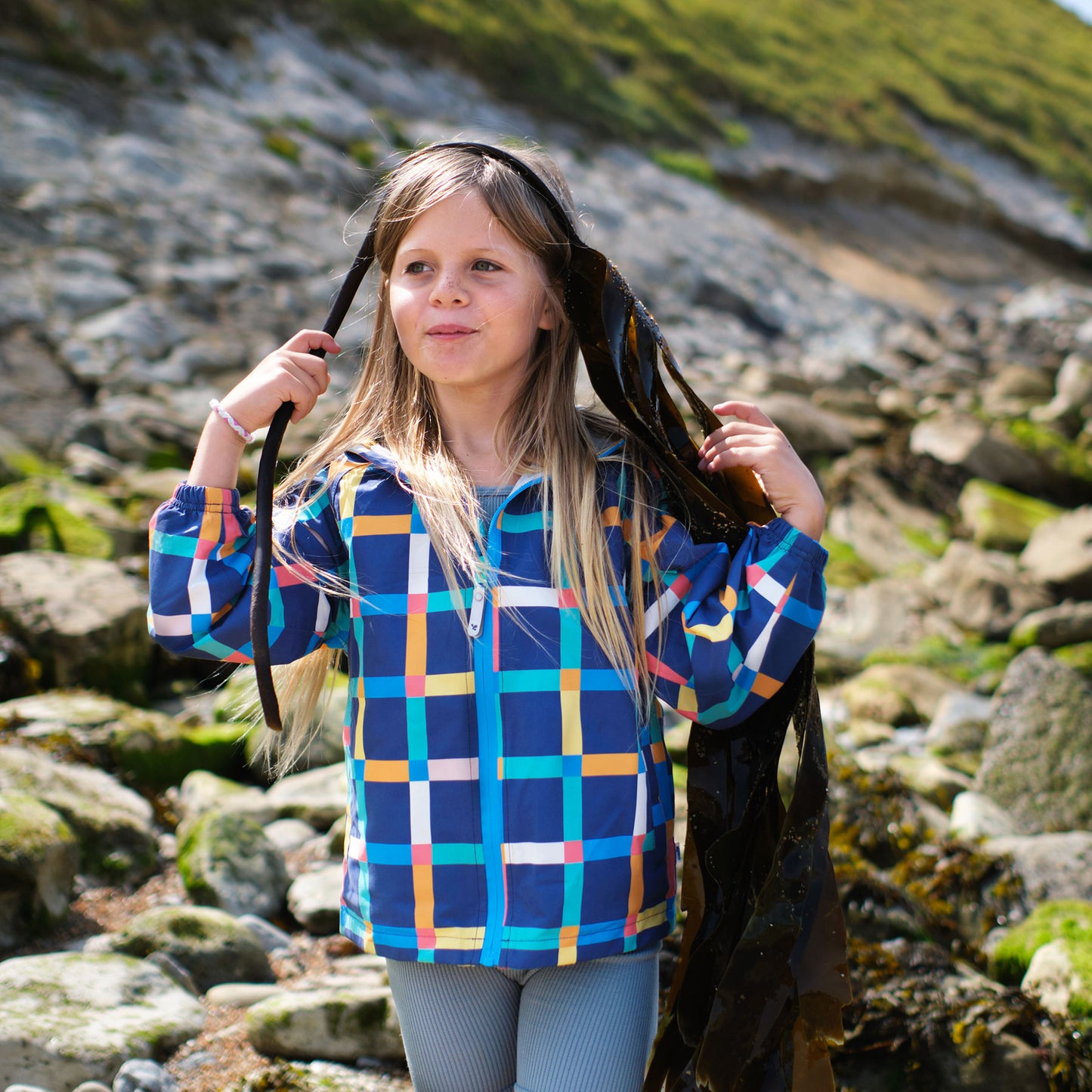 A girl on a rocky beach wearing an EcoLight Lightweight Waterproof Jacket Multi Check, holding seaweed near a grassy hillside.