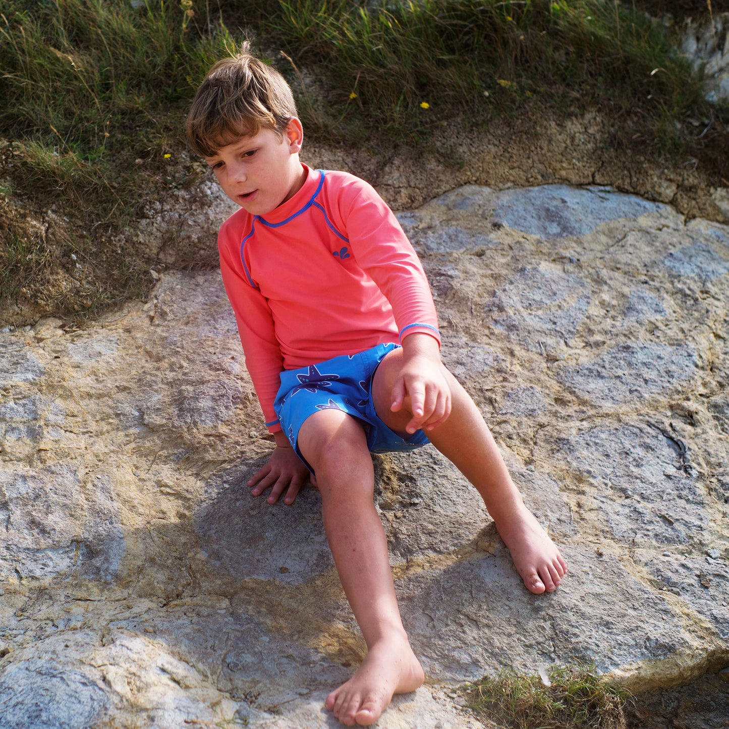 A child wearing a red UV Protective Rash Vest and blue starfish shorts sits on a rock, looking down and pointing with their right hand.