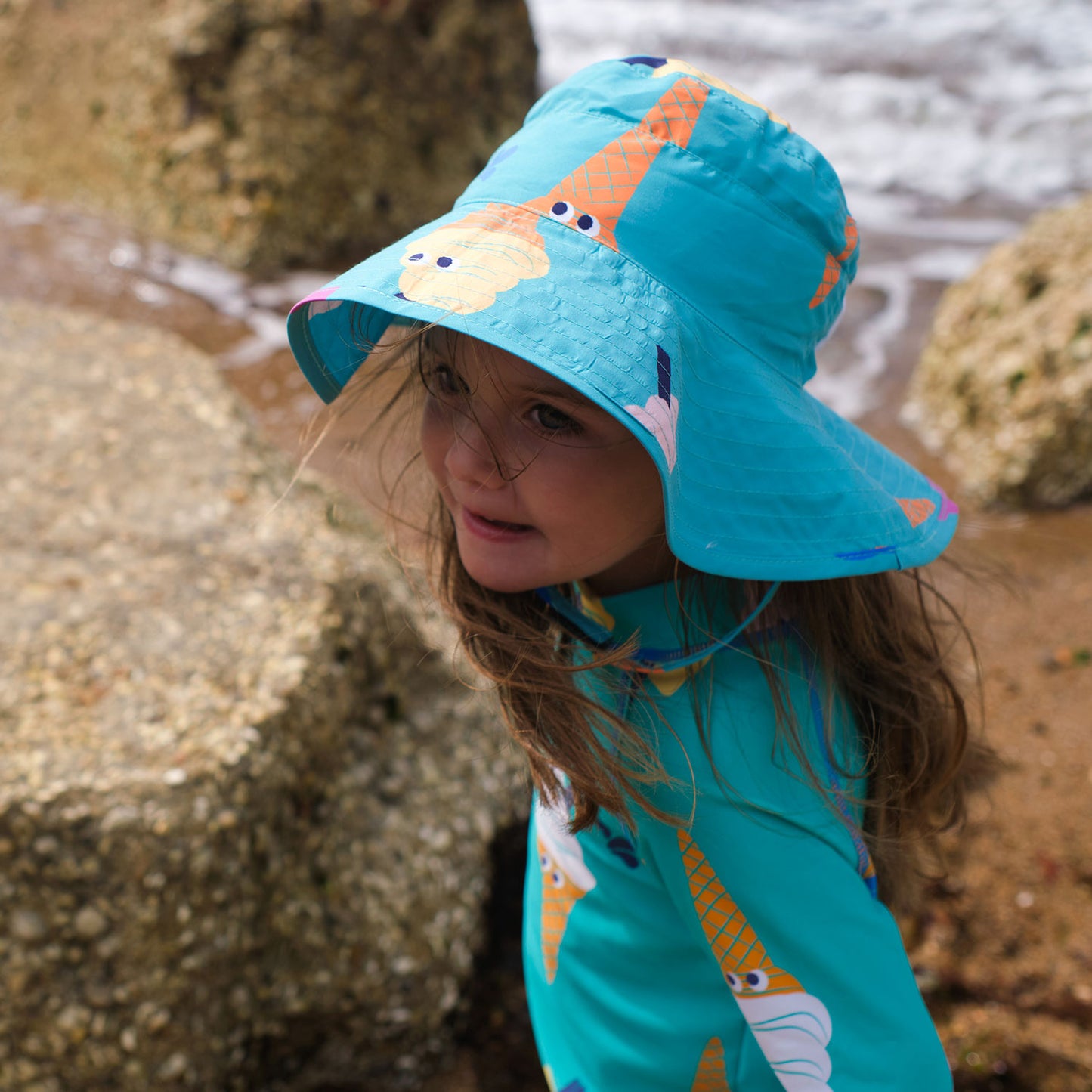 A little girl wears a 'UV Protective Sun Hat Green Ice Cream' by the sea. She is next to some rocks, looking into the distance.