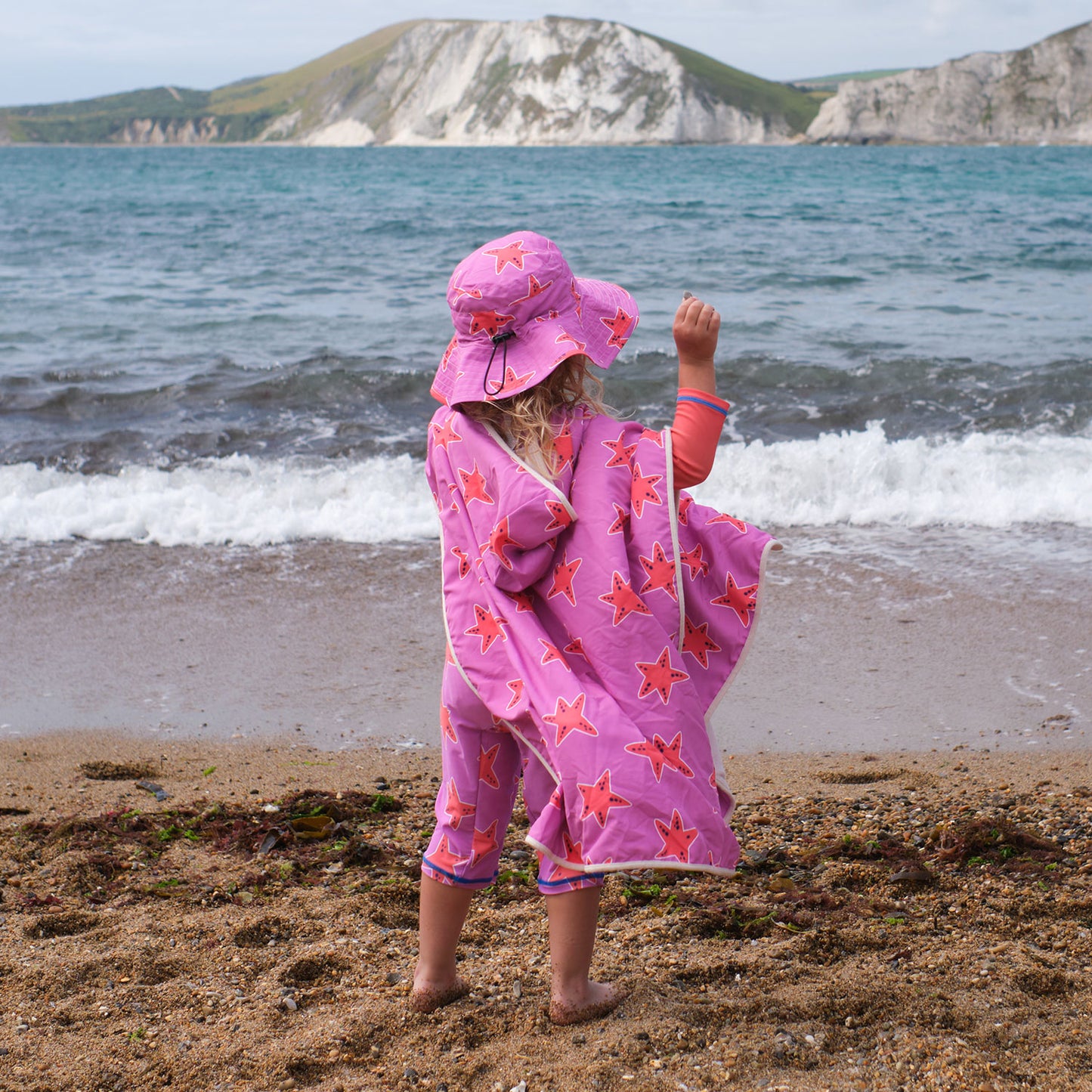 A child wearing a UV Protective Sun Hat Pink Starfish, stands on a sandy beach, back to camera, looking out at the waves of the sea.