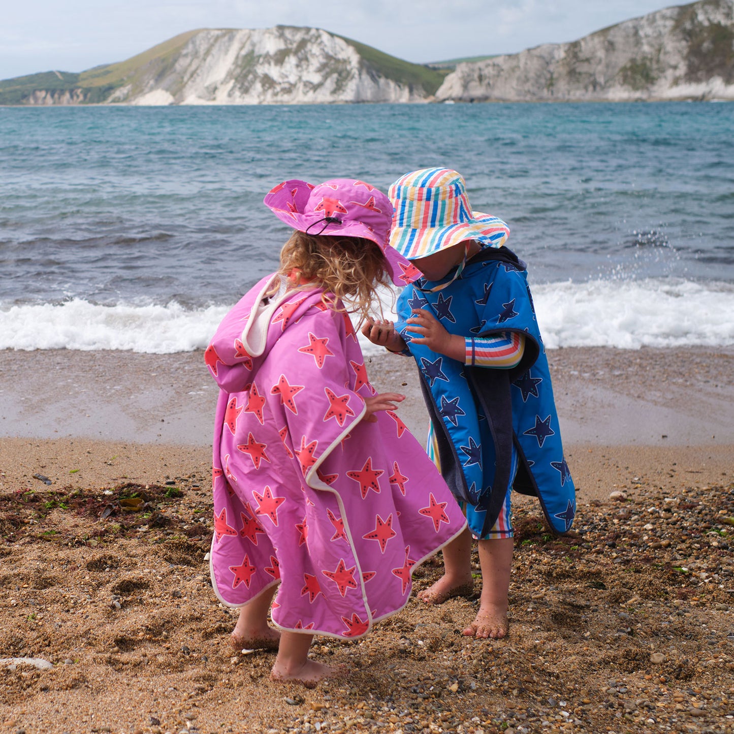 Two children wearing UV Protective Sun Hats on a beach, one pink with stars, one multicoloured stripes, standing near the sea.