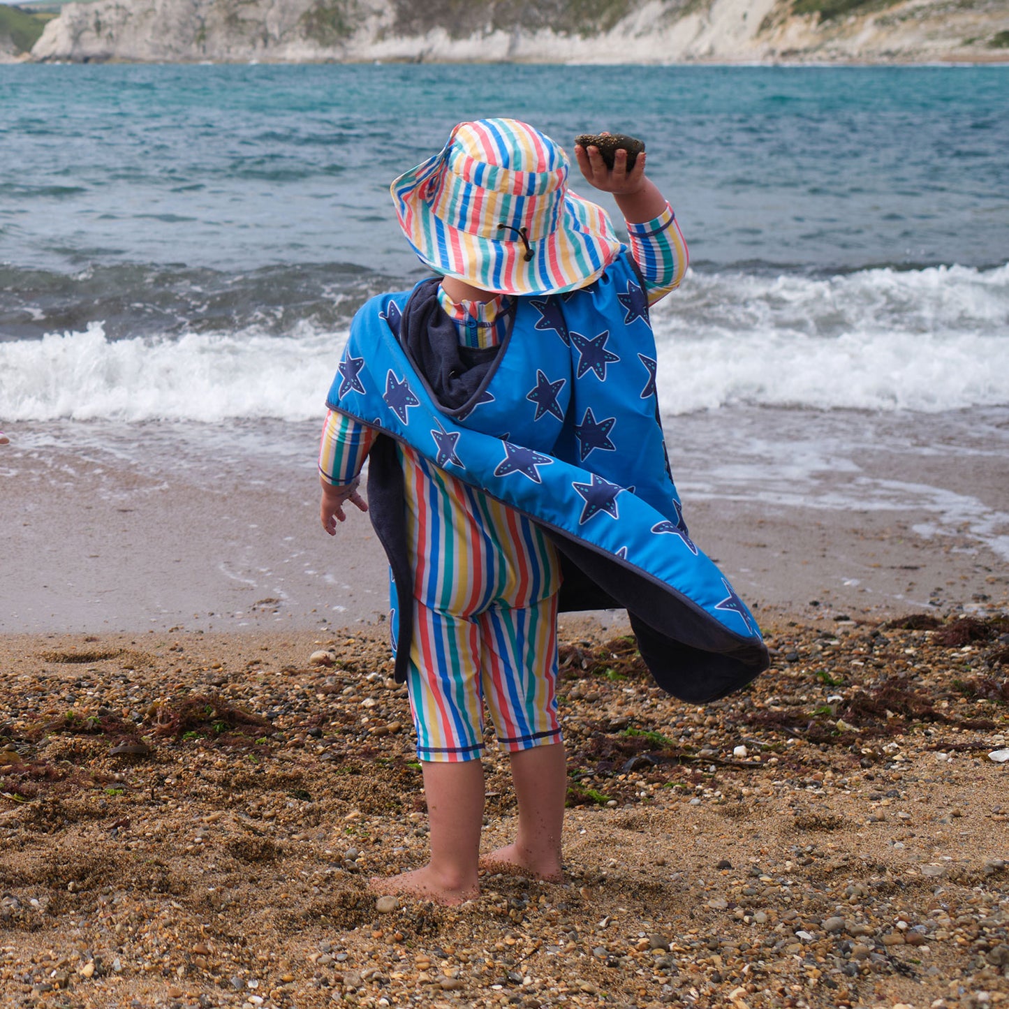 A child on a beach wearing a UV Protective Sun Hat Multi Stripe, holding a stone, with sea and cliffs in the background.