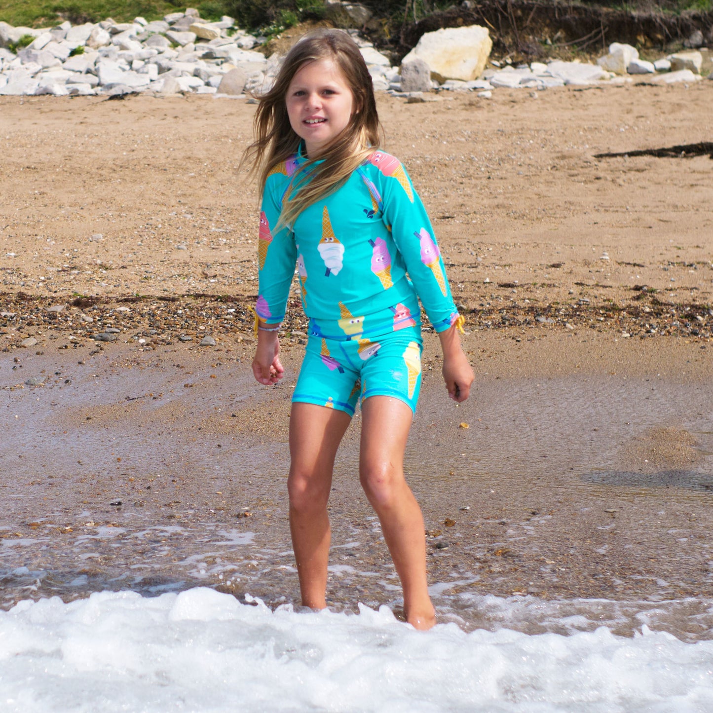 A young girl stands on a sandy beach in an Ice Cream UV Protective Surf Set, enjoying the sea breeze and waves.