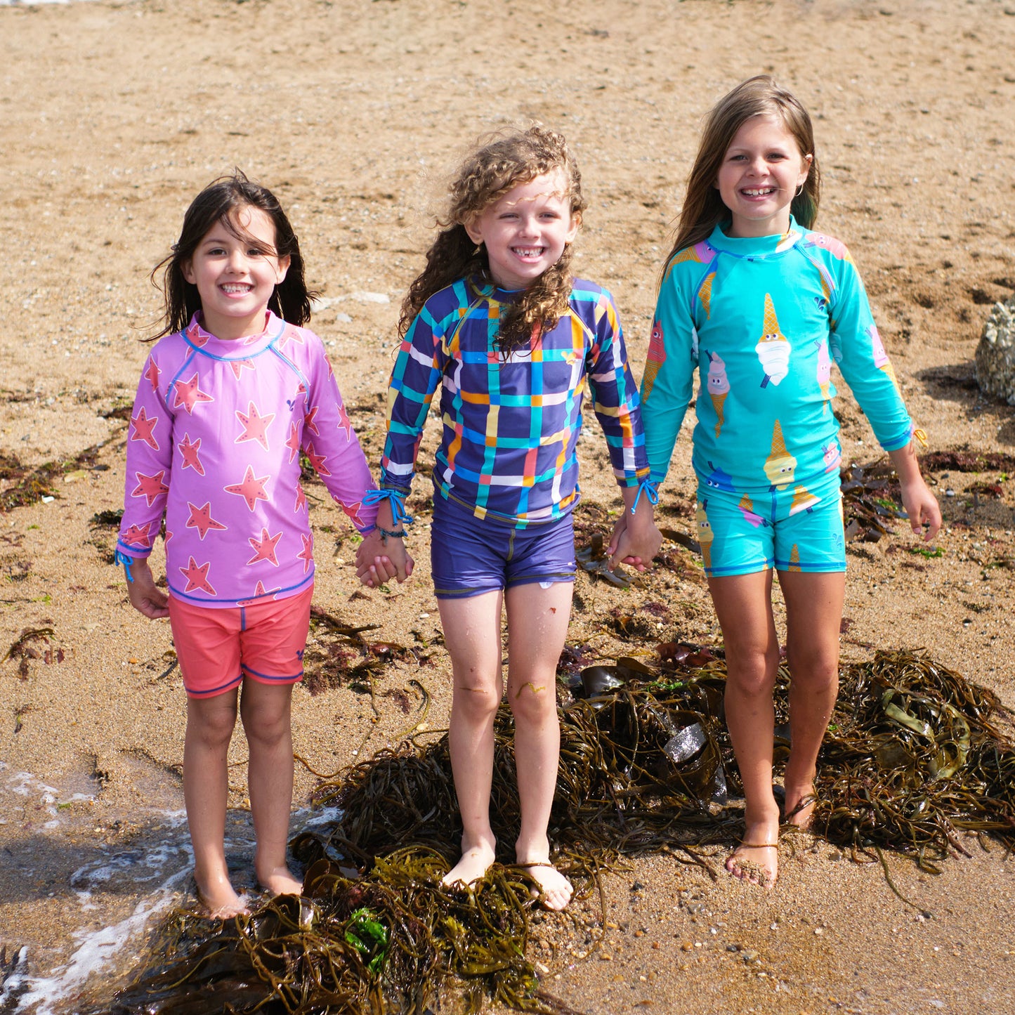 Three girls in UV Protective Surf Sets Multi Check, starfish and ice cream designs stand on a beach holding hands, smiling at the camera.