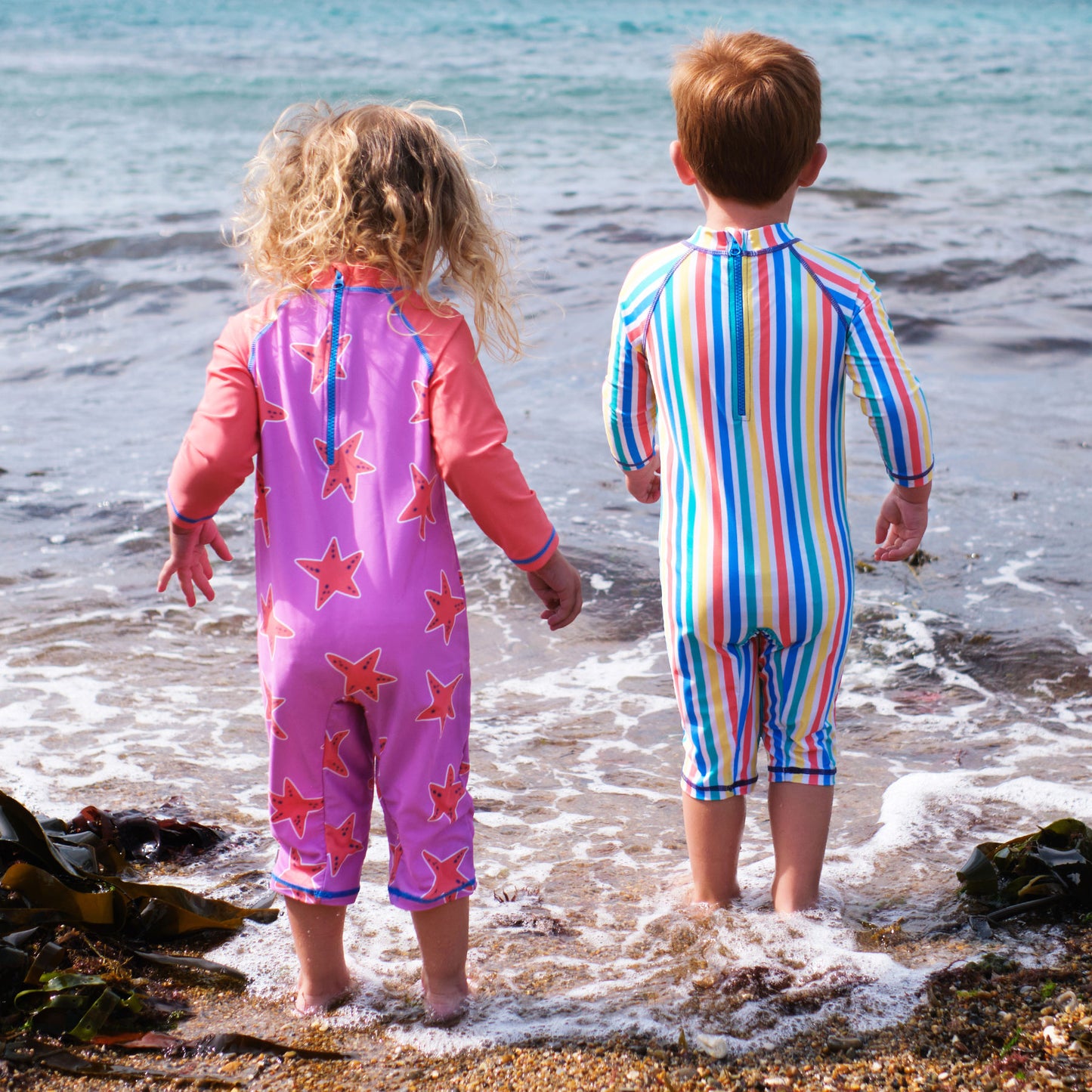 Two young children wearing UV Protective Surf Suits stand at the beach, looking out to sea, paddling in the water.