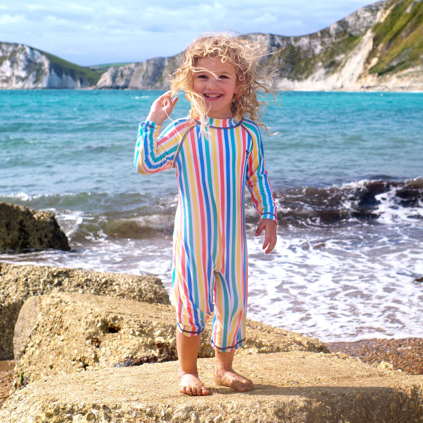 A child wearing a UV Protective Surf Suit Multi Stripe stands on rocks at the beach with the sea and cliffs in the background.