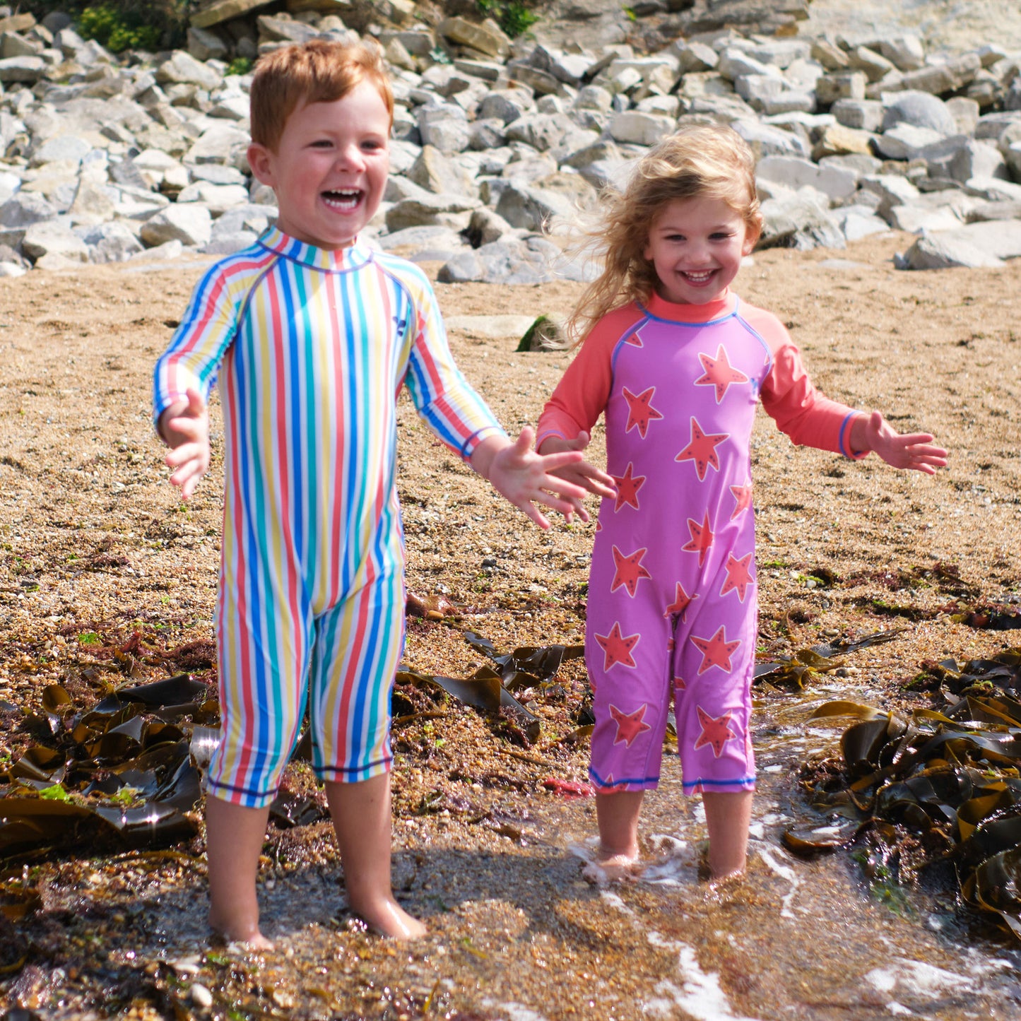 Two children in UV Protective Surf Suits Multi Stripe play in the sea, smiling with arms outstretched on a sunny day.