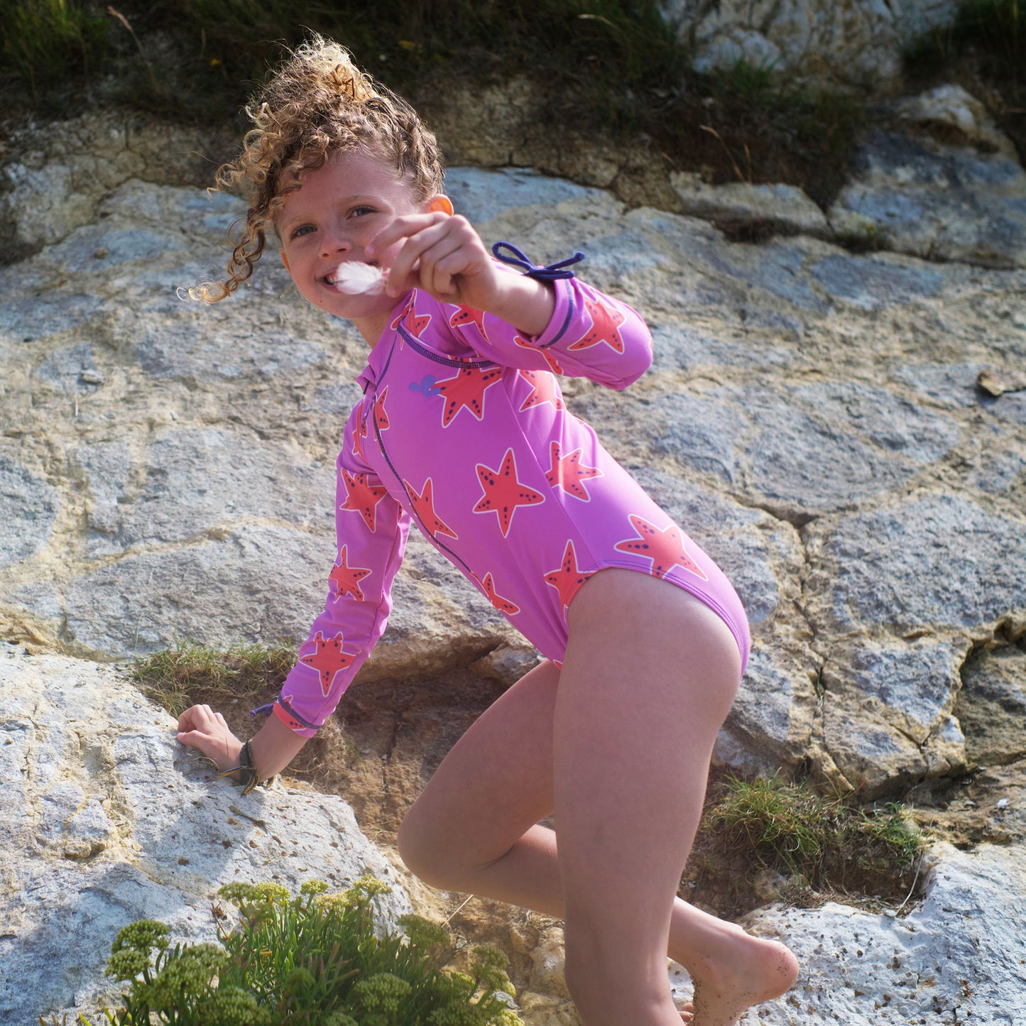 A young girl climbing a rock face wearing a UV Protective Long Sleeve Swimsuit Pink Starfish while holding a feather.