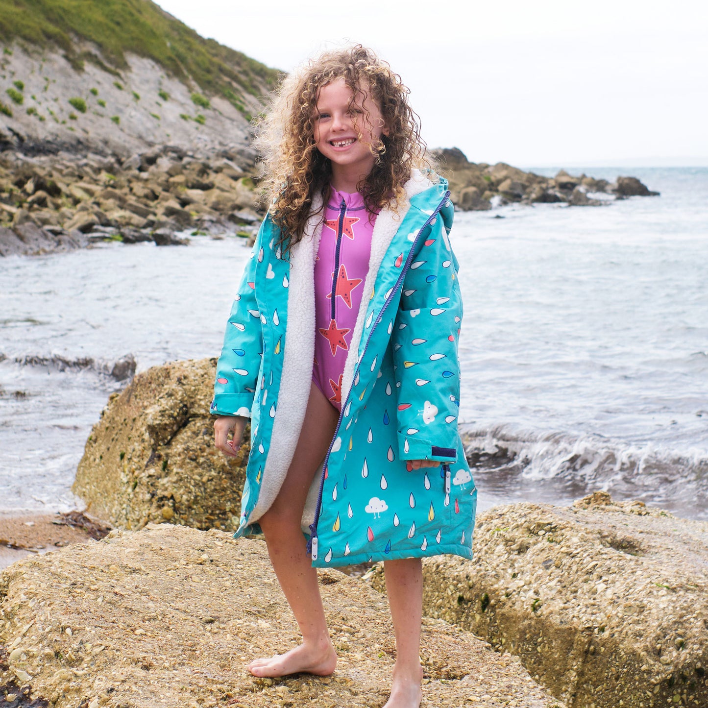 A girl on a beach wearing a Waterproof Changing Robe Green Raindrops. She is stood on a rock near the sea with a cliff behind her.