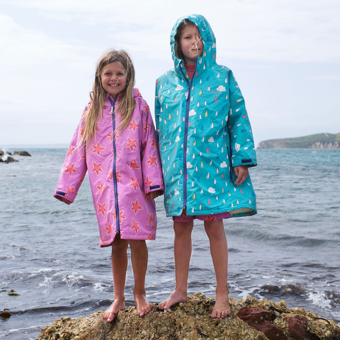 Two girls stand on a rock near the sea wearing Waterproof Changing Robes. One pink starfish robe, one blue rain cloud robe.