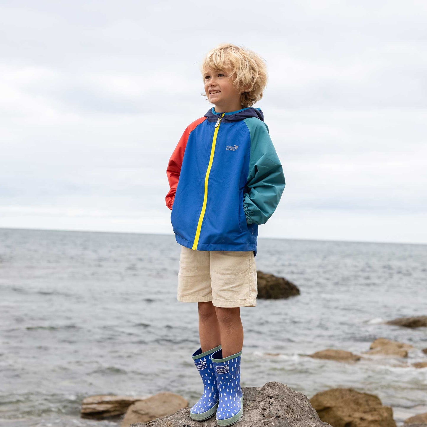 Young boy on rocky shore wearing EcoLight Lightweight Waterproof Jacket, showcasing its durable, waterproof design with taped seams and elasticated cuffs, ideal for adventurous outings.