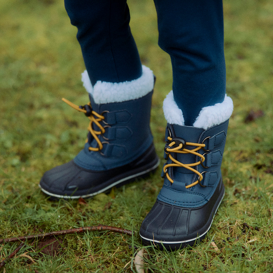 A pair of Blue Snow Boots being worn on grass. The boots are blue and black with yellow laces and a white fur lining.