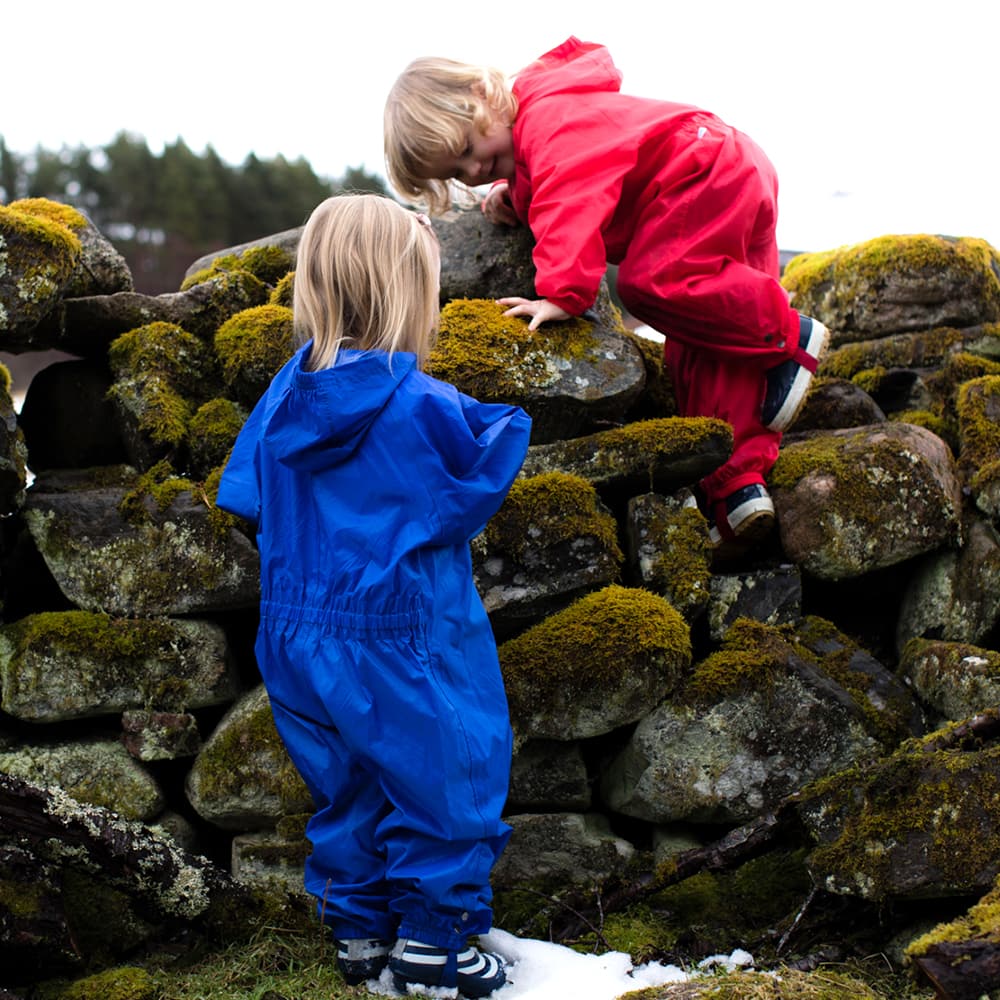 Two kids in waterproof suits play on a mossy stone wall. One wears an Originals Waterproof Puddle Suit Blue. A playful outdoor scene.
