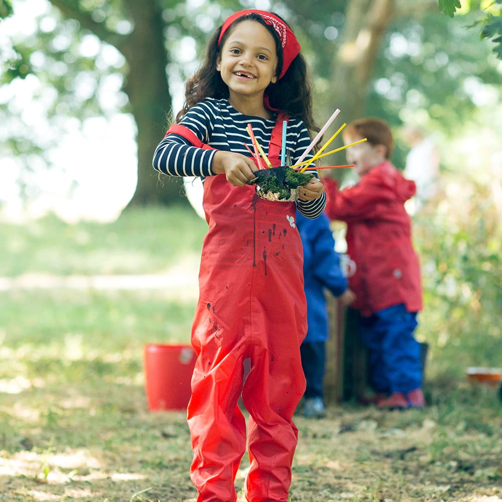 A girl wearing 'Originals Waterproof Dungarees Red', holding a small bowl of moss with coloured straws. Others play in background.
