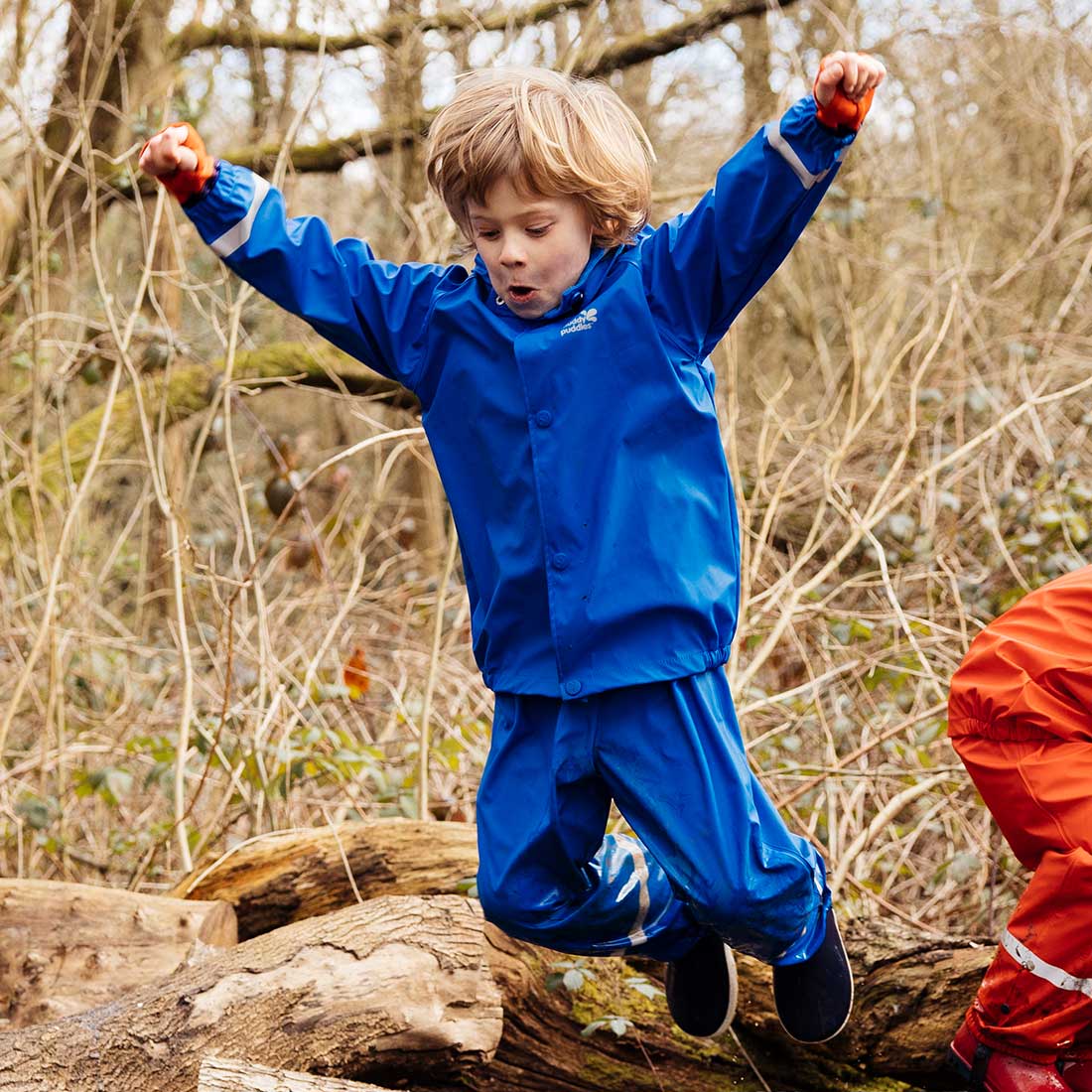 A boy jumping in the woods wearing a Rainy Day Waterproof Jacket and Dungarees Set Blue, appearing to have a great time.