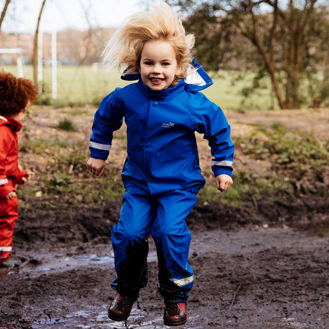A child joyfully jumping in muddy puddles in a blue Rainy Day Waterproof Jacket and Dungarees Set Blue on a cloudy day.
