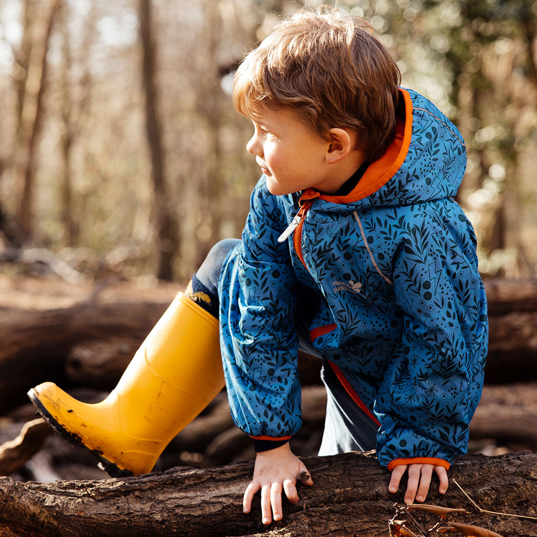 A young boy in Classic Wellies Yellow climbs a log in a forest. He's wearing a blue patterned jacket, and the sunlight dapples the scene.