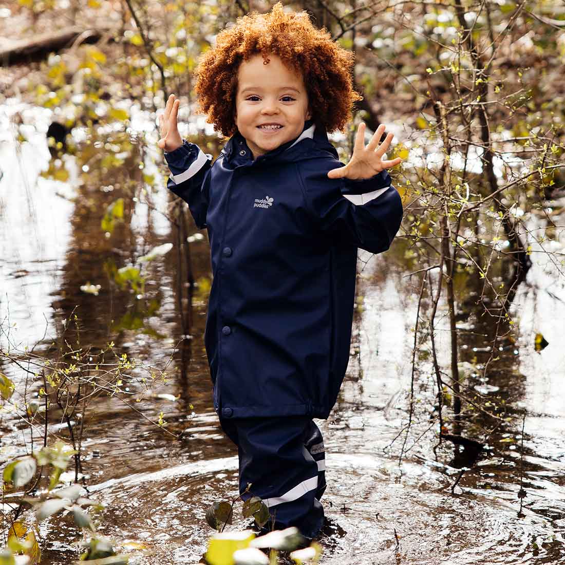 A young child in a Rainy Day Waterproof Jacket and Trousers Set Navy, standing in a puddle, smiling with arms raised amongst trees.