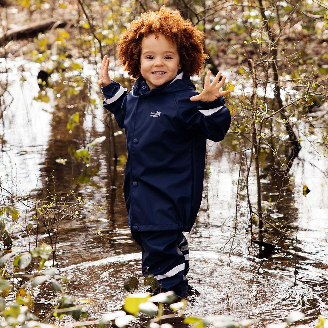 A young child stands in a watery woodland wearing a Rainy Day Waterproof Jacket and Dungarees Set Navy, smiling with hands raised.