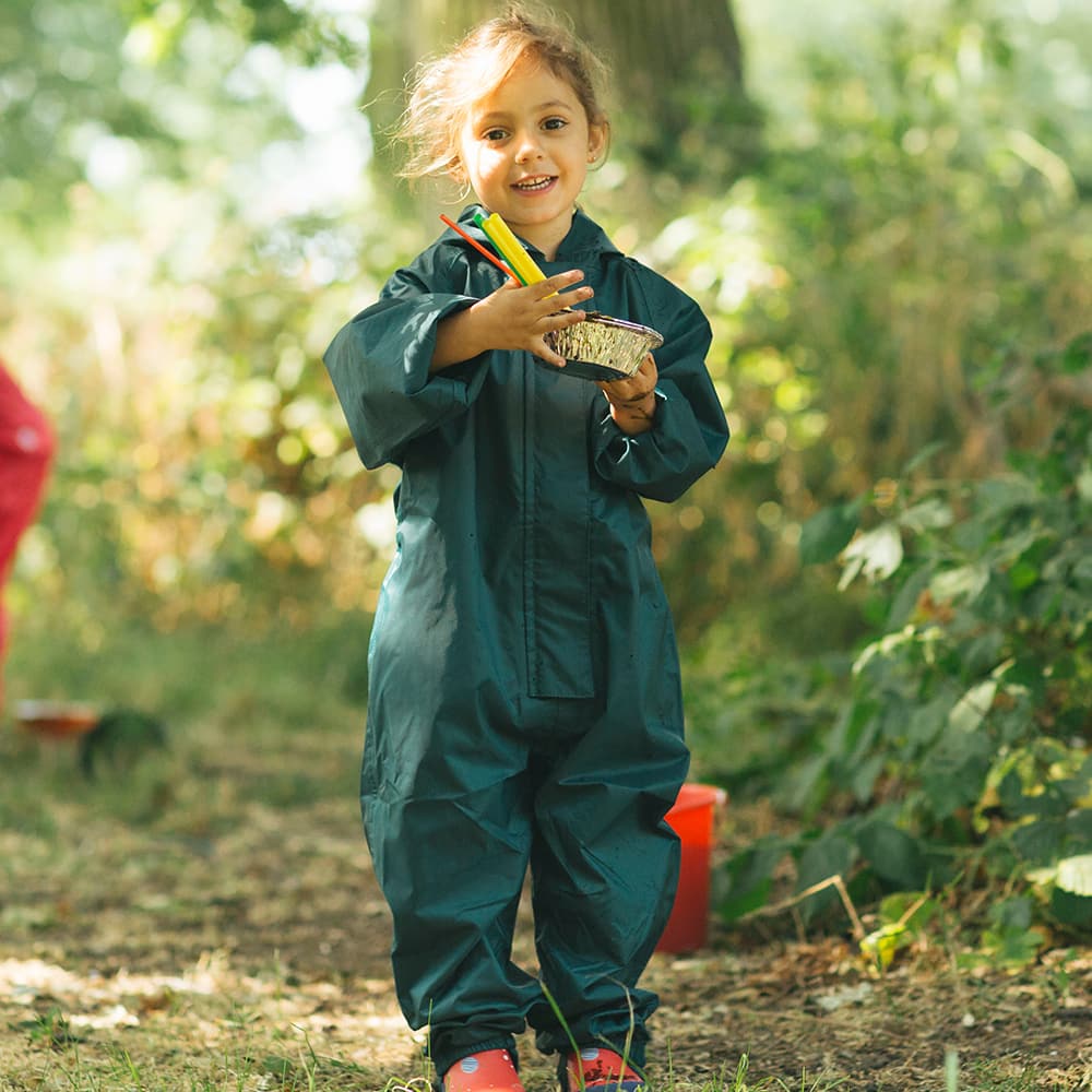 A girl in a green Originals Waterproof Puddle Suit stands outside, holding crayons and a foil dish, smiling at the camera.
