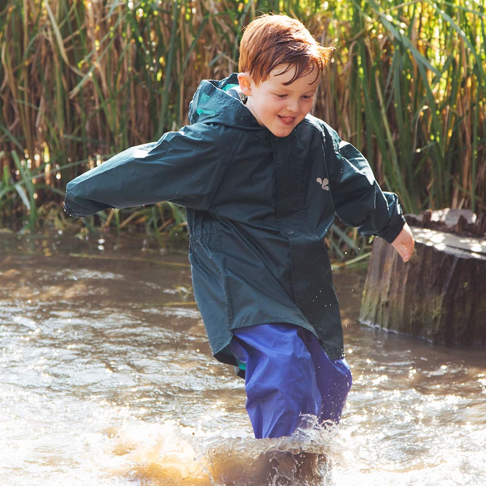 A happy red-haired child wearing the Originals Waterproof Jacket Green splashing through water outdoors on a sunny day.