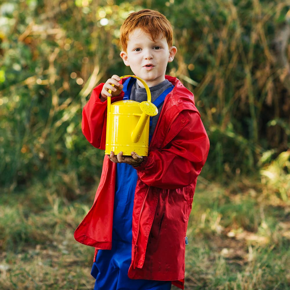 A young ginger haired boy holds a yellow watering can whilst wearing a red Originals Waterproof Jacket in a garden.