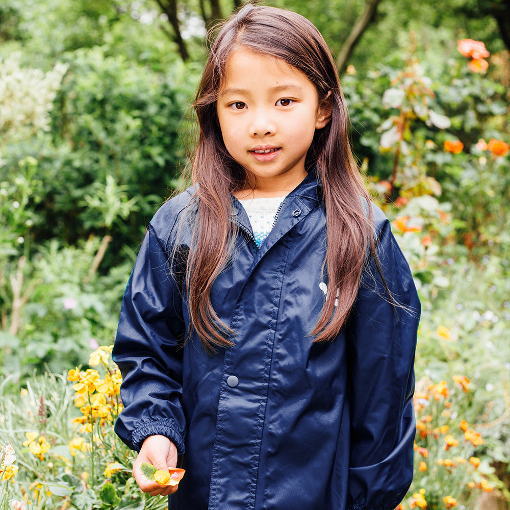 A young girl stands in a garden, wearing a navy Originals Waterproof Jacket, surrounded by flowers.