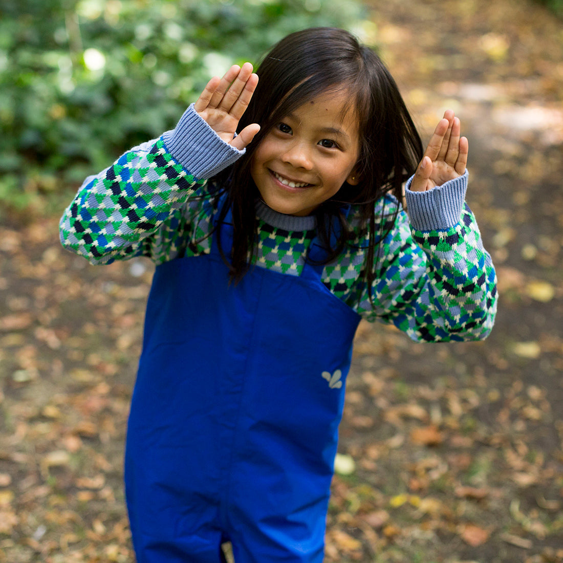 A child smiles, wearing Originals Waterproof Dungarees Blue with a patterned jumper. The child has their hands up and is outside in a garden.