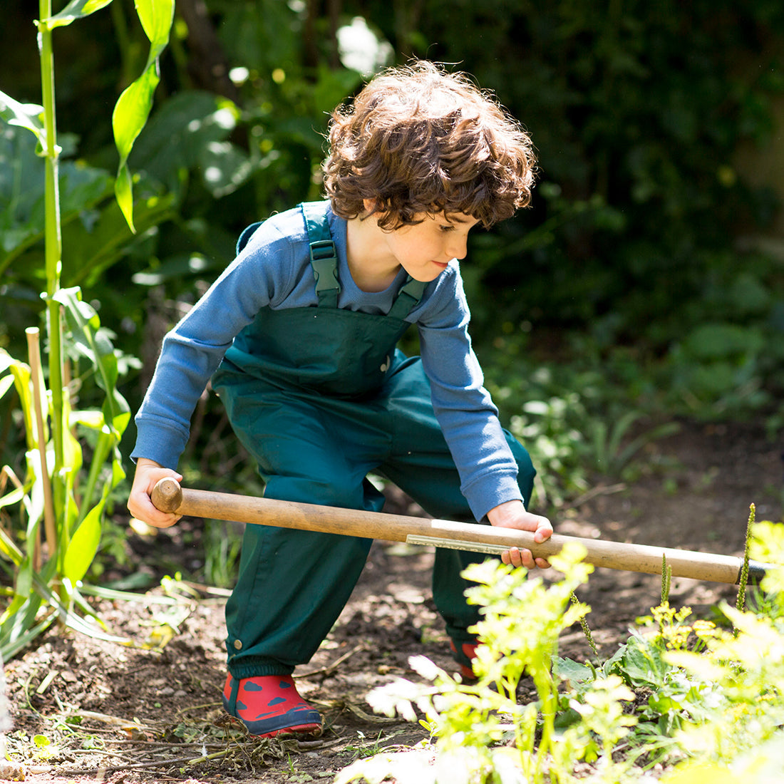 A boy in a garden wearing green Originals Waterproof Dungarees, using a garden hoe. He has curly brown hair, and is wearing red wellington boots.