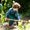 A boy in a garden wearing green Originals Waterproof Dungarees, using a garden hoe. He has curly brown hair, and is wearing red wellington boots.