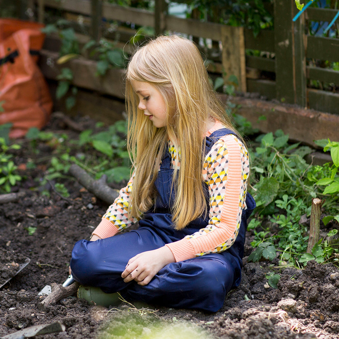 A girl is in a garden wearing Originals Waterproof Dungarees in navy. She is digging and looking at the earth with leaves around her.