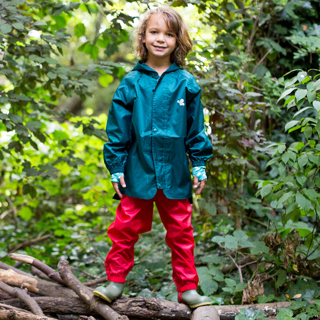 A boy standing on a log wearing red Originals Waterproof Trousers, a green waterproof jacket and boots in a forest setting.