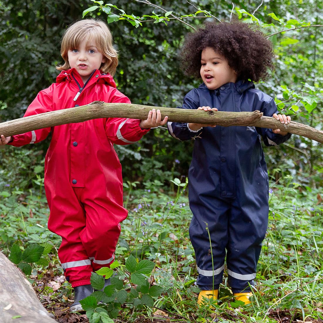 Two children, one is a boy, outdoors in Puddleflex Waterproof Fleece Lined Puddle Suits, holding a log. One suit is red, the other navy blue.