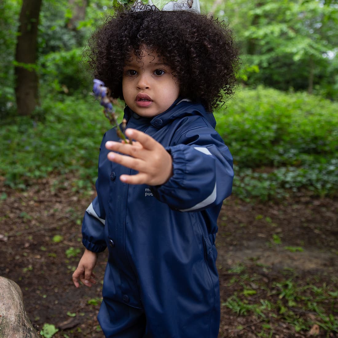 A young child wearing a Puddleflex Waterproof Fleece Lined Puddle Suit in Navy, holding a flower in a woodland setting.