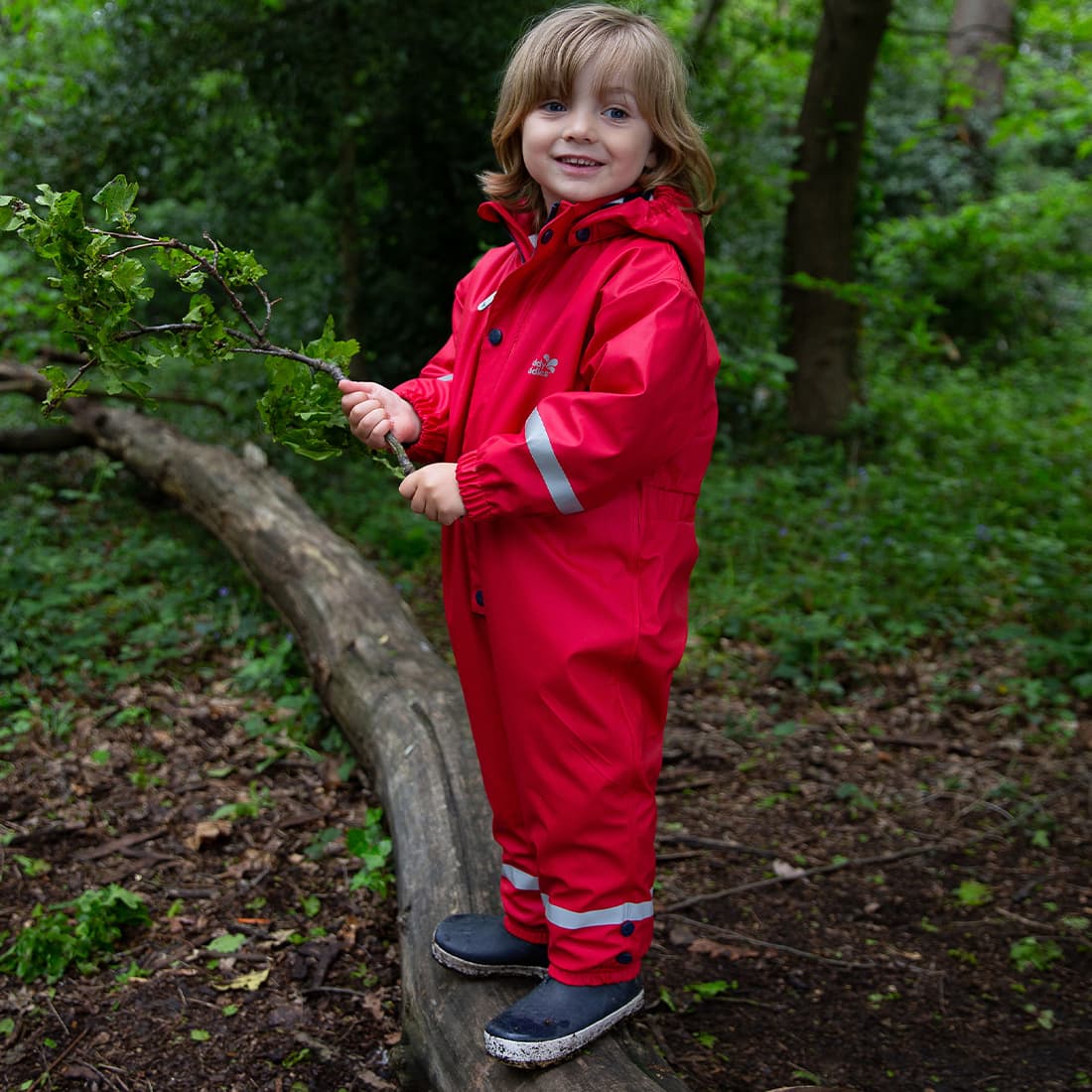 A child stands on a log in a forest, wearing a red Puddleflex Waterproof Fleece Lined Puddle Suit while holding a leafy branch.